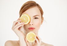 Young beautiful woman with clean healthy skin looking at camera hiding eye behind lemon slice over white background.