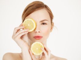 Young beautiful woman with clean healthy skin looking at camera hiding eye behind lemon slice over white background.